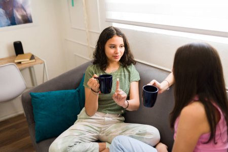 Photo for Hispanic teen girl drinking a cup of tea or chocolate while talking with her best friend and having a fun time at home - Royalty Free Image