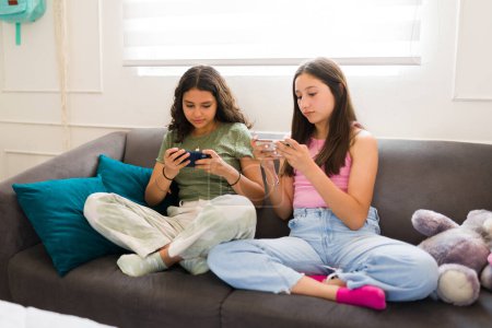 Photo for Beautiful teenagers and best friends sitting together on the couch and texting on their smartphones while hanging out at home - Royalty Free Image