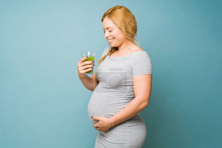 Téléchargez les photos : Mignon caucasien enceinte femme rester en bonne santé en buvant vert jus et sourire dans un studio - en image libre de droit