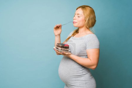 Photo for Beautiful pregnant woman giving into her cravings and enjoying a big piece of cake in a studio - Royalty Free Image