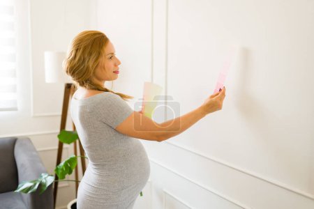 Photo for Cute pregnant woman holding color swatches and trying to choose the best color for painting the baby nursery room - Royalty Free Image