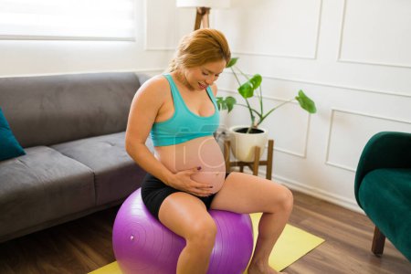 Photo for Beauitful pregnant woman touching her belly while sitting on a stabilty ball and exercising at home - Royalty Free Image