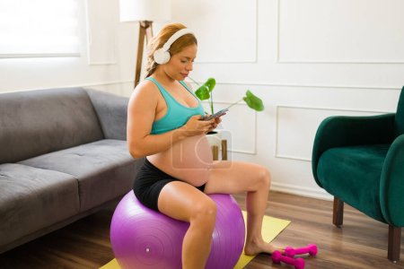 Photo for Expectant mother in her 30s wearing headphones and choosing the right playlist on her smartphone while working out at home - Royalty Free Image
