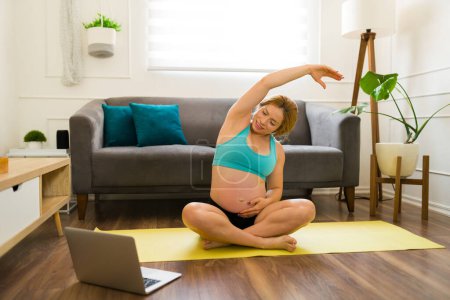Photo for Pregnant woman doing some stretching exercises and warming up while doing an online course from home - Royalty Free Image