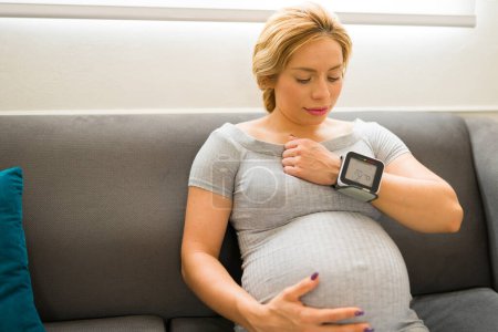 Téléchargez les photos : Femme enceinte vérifiant sa pression artérielle avec un moniteur de fréquence cardiaque au poignet à la maison - en image libre de droit