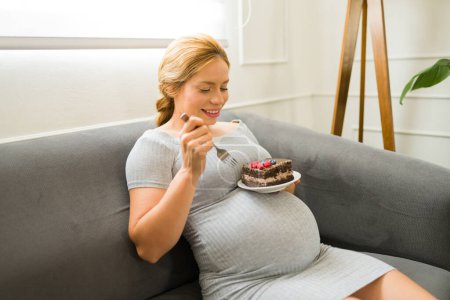 Foto de Mujer atractiva disfrutando de su embarazo y comiendo una rebanada grande y deliciosa de pastel como uno de sus antojos - Imagen libre de derechos