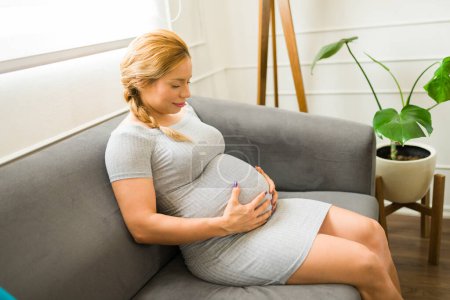 Foto de Retrato de una linda mujer embarazada sentada en un sofá en casa y tocándose el vientre - Imagen libre de derechos