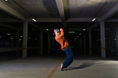 Téléchargez les photos : Funky street male dancer danse sur la musique urbaine dans un parking sombre et profiter d'une chorégraphie freestyle - en image libre de droit