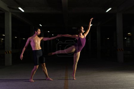 Photo for Beautiful young woman in a leotard dancing classic ballet with an hispanic artistic man at night in the parking lot - Royalty Free Image