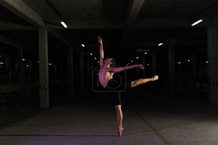 Foto de Atractiva bailarina latina bailando por la noche en un estacionamiento oscuro y haciendo una pose arabesca de ballet clásico - Imagen libre de derechos