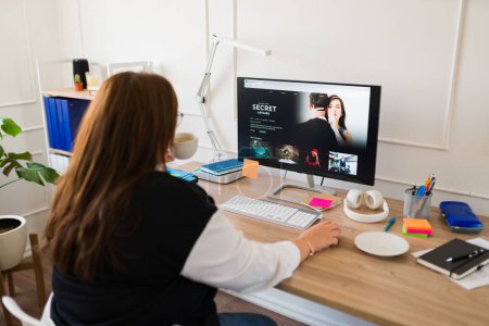Photo for Professional woman seen from behind drinking coffee at her office desk and watching movies or series during business hours - Royalty Free Image