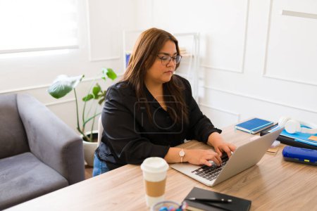 Téléchargez les photos : Femme d'affaires professionnelle et gestionnaire tapant sur l'ordinateur portable et buvant du café tout en travaillant à son bureau - en image libre de droit