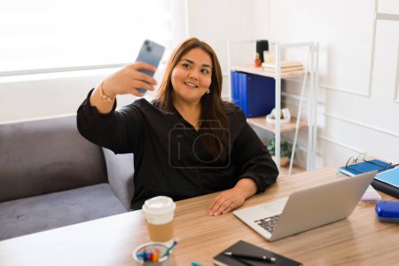 Téléchargez les photos : Attrayant plus taille femme travaillant comme gestionnaire ou patron tout en prenant un selfie avec un smartphone dans son bureau - en image libre de droit