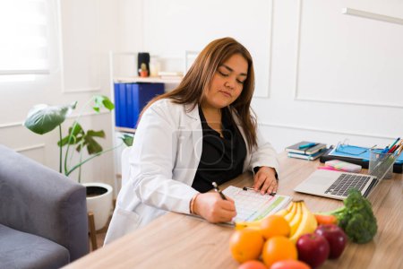 Téléchargez les photos : Latin plus taille femme nutritionniste écrit dans son bureau et prêt pour un patient de parler de saine alimentation et l'alimentation - en image libre de droit