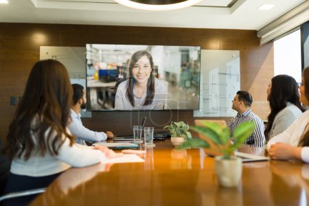 Téléchargez les photos : Femme d'affaires vue sur grand écran de télévision ayant une vidéoconférence avec un groupe de gens d'affaires assis dans une salle de réunion de bureau moderne - en image libre de droit