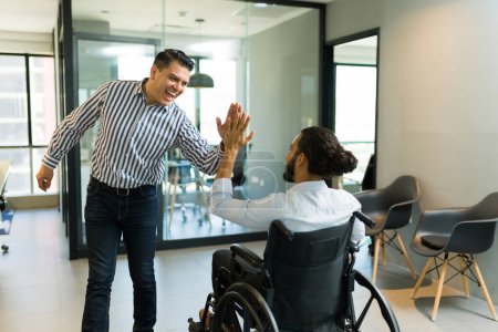 Téléchargez les photos : Joyeux milieu adulte homme d'affaires donnant haute cinq à un collègue handicapé en fauteuil roulant dans le bureau - en image libre de droit