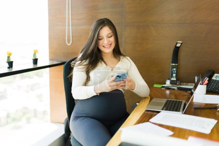 Téléchargez les photos : Femme d'affaires enceinte et souriante assise à son bureau textant sur son téléphone portable au bureau - en image libre de droit