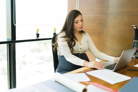 Téléchargez les photos : Femme d'affaires enceinte assise à son bureau lisant un e-mail sur son ordinateur portable pendant qu'elle travaillait au bureau - en image libre de droit