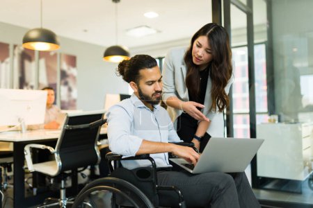 Téléchargez les photos : Femme d'affaires asiatique discutant du travail avec un homme d'affaires handicapé assis sur un fauteuil roulant en utilisant un ordinateur portable au bureau - en image libre de droit