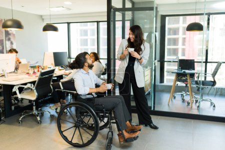 Photo for Businessman with disability on wheelchair having coffee and talking with female colleague standing by in office during coffee break - Royalty Free Image