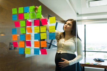 Téléchargez les photos : Femme d'affaires enceinte mettant des notes collantes colorées sur le mur de verre au bureau - en image libre de droit