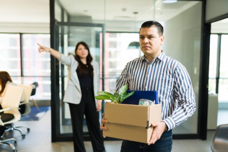Photo for Depressed mid adult male employee holding box of belongings being dismissed by angry boss in office - Royalty Free Image