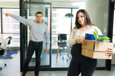 Photo for Upset pregnant female employee holding box of belongings being fired by angry boss in office - Royalty Free Image