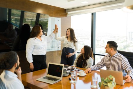 Téléchargez les photos : Femme enceinte donnant haute cinq à plus taille femme d'affaires avec l'équipe assise à la table de salle de réunion dans le bureau moderne - en image libre de droit