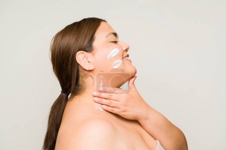 Foto de Perfil de una mujer grande obesa feliz que se pone crema hidratante y sonríe mientras cuida su piel corporal - Imagen libre de derechos
