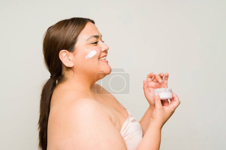 Téléchargez les photos : Vue latérale d'une femme obèse heureuse souriante tout en hydratant la crème et en utilisant des produits de beauté pour la peau - en image libre de droit