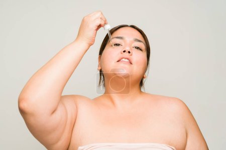 Téléchargez les photos : Grande femme obèse prévenant le vieillissement et les rides sur son visage lors de l'utilisation de sérum d'acide hyaluronique et de produits de soins de la peau - en image libre de droit