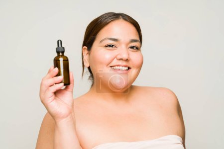 Foto de Mujer latina gorda alegre sonriendo mientras muestra una botella de producto de ácido hialurónico mientras usa productos para el cuidado de la piel de belleza - Imagen libre de derechos
