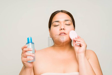 Foto de Mujer grande latina con sobrepeso y hombros desnudos usando productos de belleza que eliminan el algodón y el maquillaje limpiando su piel - Imagen libre de derechos