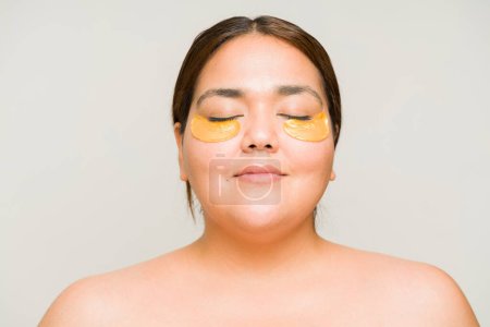 Photo for Happy attractive large woman with bare shoulders smiling while using eye patches and face masks during her skin care routine - Royalty Free Image