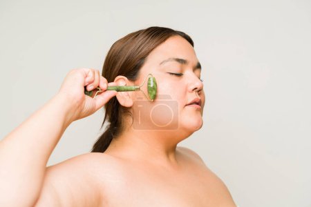 Foto de Mujer con sobrepeso segura de sí misma usando un rodillo de jade para una piel suave y hermosa y usando crema hidratante con hombros desnudos - Imagen libre de derechos