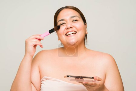 Téléchargez les photos : Enthousiaste femme heureuse en surpoids avec les épaules nues en utilisant une brosse de maquillage et se préparer à sortir tout en riant - en image libre de droit