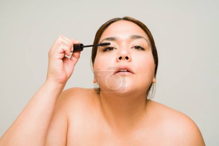 Photo for Big plus size woman looking at the camera while putting on mascara and using makeup products looking beautiful - Royalty Free Image