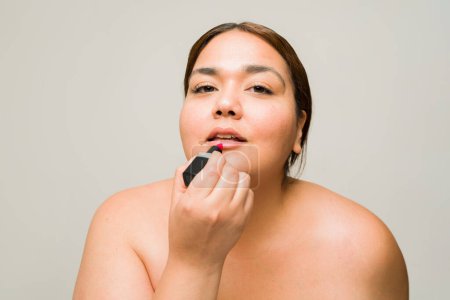 Foto de Hermosa mujer latina gorda usando lápiz labial y maquillándose después de hacer su rutina de cuidado de la piel aislada en un fondo de estudio - Imagen libre de derechos