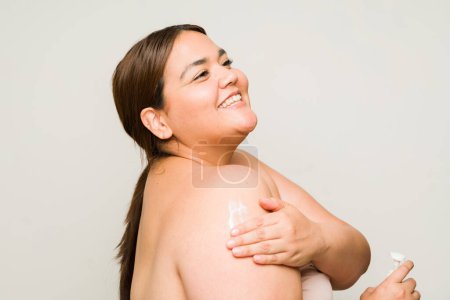 Foto de Feliz hermosa mujer de talla grande usando crema hidratante en su cuerpo mientras usa productos de cuidado de belleza - Imagen libre de derechos