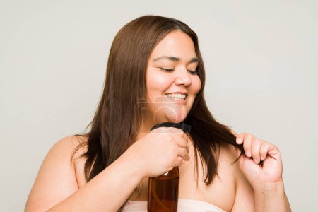 Foto de Mujer hispana con sobrepeso sonriendo usando aceite en aerosol en sus puntas abiertas y cuidando su hermoso cabello - Imagen libre de derechos
