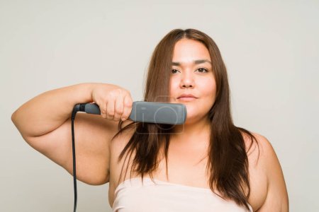 Photo for Plus size hispanic woman doing a new hairstyle and using a straightening iron while looking at the camera isolated in a studio background - Royalty Free Image