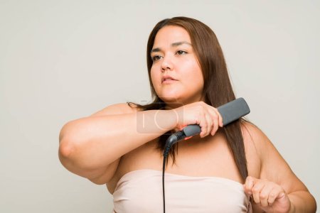 Foto de Atractiva mujer latina gorda de unos 30 años usando una plancha enderezadora y preparándose para salir aislada en un estudio - Imagen libre de derechos