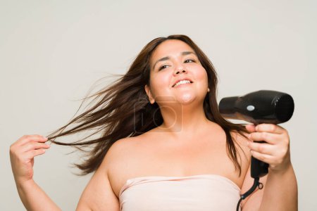 Photo for Cheerful relaxed obese woman smiling while blowing her hair using a blow dryer after showering with bare shoulders - Royalty Free Image
