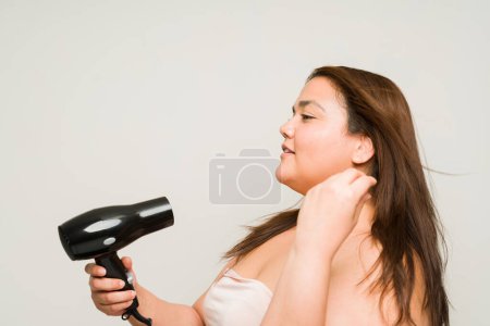 Téléchargez les photos : Vue latérale d'une grosse femme en surpoids séchant ses cheveux à l'aide d'un sèche-cheveux se préparant à sortir après la douche en souriant - en image libre de droit