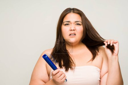 Photo for Sad upset plus size woman in her 30s with damaged dry hair suffering from split ends while brushing her hair making eye contact - Royalty Free Image