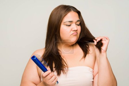 Foto de Mujer enojada con sobrepeso con el cabello seco dañado y puntas abiertas cepillándose el pelo y sintiéndose triste - Imagen libre de derechos
