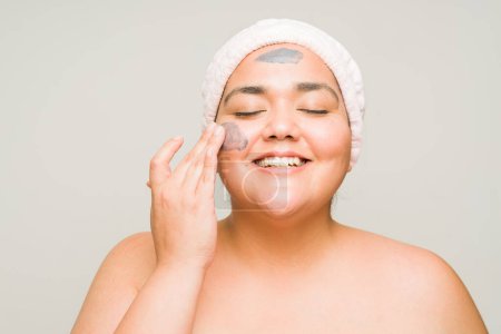 Téléchargez les photos : Heureuse femme hispanique de graisse souriante portant un masque de boue et souriant en utilisant des produits de soins de la peau isolés dans un arrière-plan de studio - en image libre de droit