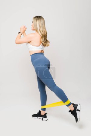 Photo for Athletic fit girl training while working out her legs and booty with resistance band exercises against a white studio background - Royalty Free Image