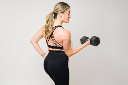 Foto de Hermosa mujer de fitness levantando pesas de mancuerna y haciendo rizos de bíceps durante su entrenamiento atlético y entrenamiento visto desde atrás - Imagen libre de derechos