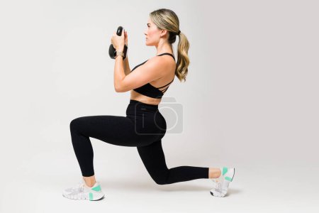 Photo for Sporty athletic woman doing bulgarian squats while using a kettlebell weight at the gym against a white studio background - Royalty Free Image